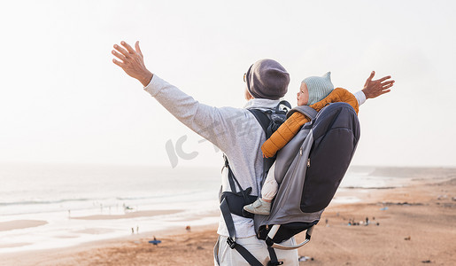年轻的父亲举起手向天空，同时在多风的沙滩上背着他的婴儿男婴背包享受纯净的大自然。