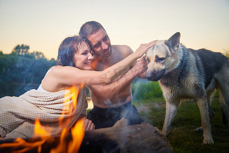 快乐的湿夫妇放松，在日落的夏日阳光明媚的夜晚，在大自然中露营，与大狗在火边玩乐和拥抱。
