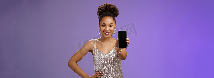 时髦优雅的非洲裔美国女性穿着闪闪发光的银色连衣裙，手握腰部自信的姿势，高兴地微笑着伸出手臂，展示智能手机显示屏，看看酷炫的应用程序蓝色背景