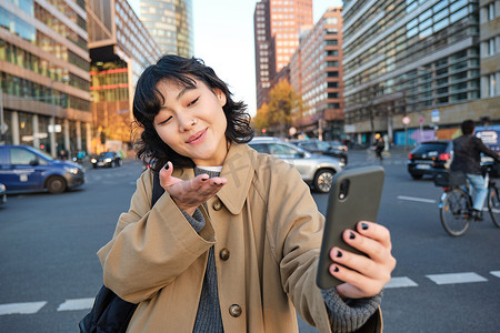 可爱的韩国女孩，游客自拍，与朋友视频聊天并向相机发送亲吻，用智能手机在城市街道上录制视频博客