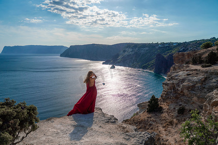 一位身穿红色长裙、头发飘逸的女孩站在海面的岩石上。