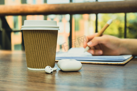 工艺回收纸杯中的咖啡变得面目全非，女人用无线耳机在纸质笔记本上写字。