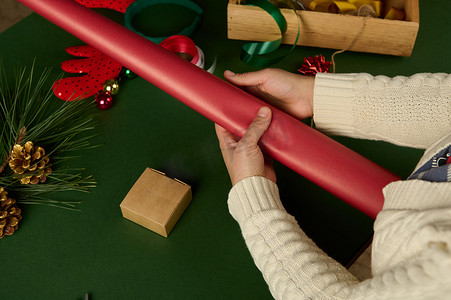 特写女人拿着红色包装礼品纸，同时在绿色背景下包装圣诞或新年礼物。