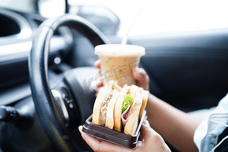 亚洲女司机拿着冰咖啡杯和三明治面包在车里吃喝，很危险，有发生事故的风险。