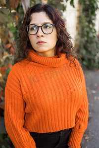 时尚橙色摄影照片_秋季肖像的沉思悲伤的年轻女子在时尚眼镜在针织时尚橙色毛衣在树叶的背景。