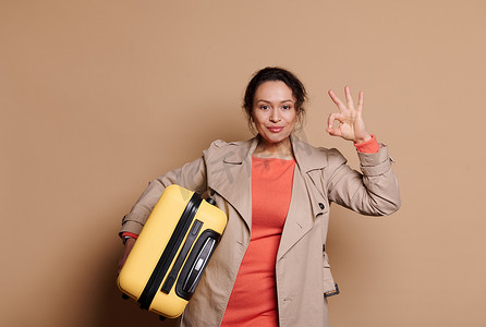可爱的少数民族女乘客提着手提箱，在米色孤立的背景上微笑着用 Ok 手势做手势