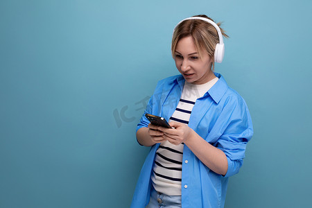 一位身着条纹毛衣和休闲衬衫、戴着白色无线耳机、面带微笑的金发年轻女子的特写照片在蓝色背景的智能手机中的应用程序中进行通信