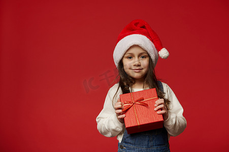 可爱的小女孩戴着圣诞老人帽，拿着带圣诞惊喜的红色礼盒，在彩色背景中突显