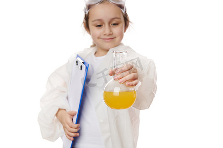 带有黄色液体化学溶液的实验室烧瓶，在模糊的学童手中，穿着带有剪贴板的实验室外套