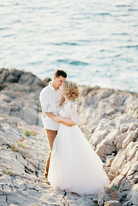 新郎在海边的岩石上拥抱穿着白色蕾丝连衣裙的新娘