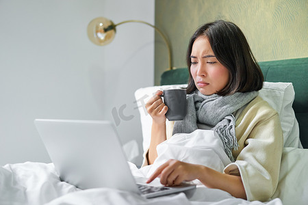 亚洲女孩看着笔记本电脑，感觉不舒服，感冒，喝热茶喉咙痛，皱着眉头看着电脑屏幕