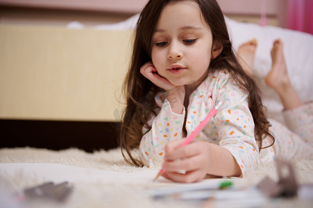 穿着睡衣的可爱小女孩躺在卧室的地毯上，专心致志地用彩色铅笔画画