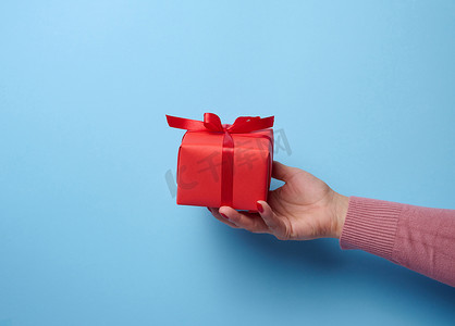 一个女人的手拿着一个蓝色背景上用丝带包裹的红色礼盒，这是一个祝贺、惊喜的概念