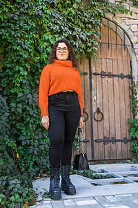 针织衫摄影照片_秋季画像是一位戴着时尚眼镜、身穿橙色针织衫的迷人年轻女子的画像，背景是树叶和大门。