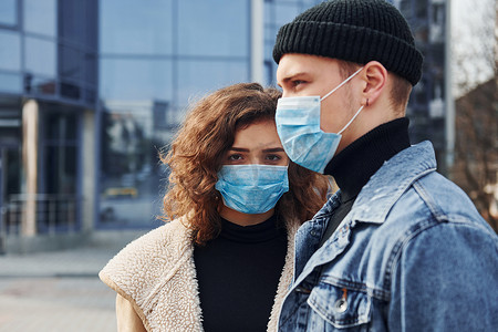 感染摄影照片_一对戴着防护面具的夫妇在检疫时间在商业大楼附近的城市户外散步。