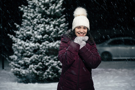 美丽的女人微笑着露齿微笑，看着相机，在雪夜里站在一棵白雪覆盖的冷杉树旁