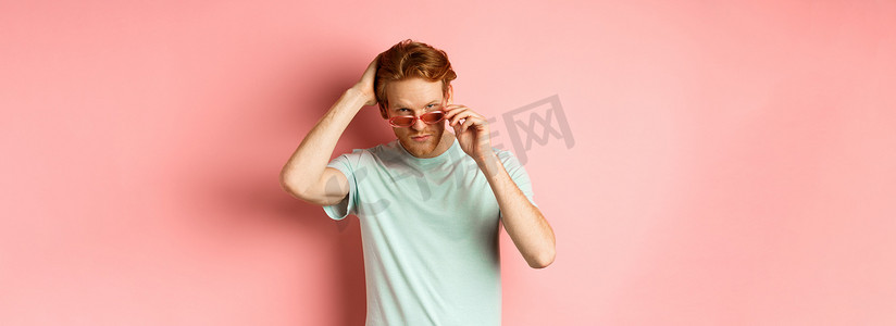 戴着墨镜的英俊年轻红发男子，用手梳理头发，站在粉红色背景上，对着镜头显得自鸣得意和自信