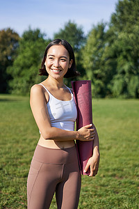 走路健身摄影照片_亚洲年轻健身女孩的垂直肖像带着橡胶垫走路瑜伽，在公园的新鲜空气中进行训练，穿运动服