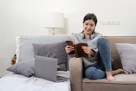 年轻漂亮的亚洲女学生坐在家里的沙发上看书，同时使用笔记本电脑进行在线学习。