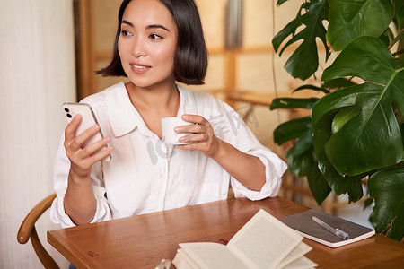 年轻的亚洲女商人坐在咖啡馆里，拿着笔记本、咖啡，看着智能手机、视频聊天，打电话给某人