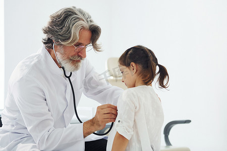 头发灰白、白大衣胡须的高级男医生在诊所用听诊器测量小女孩的心率
