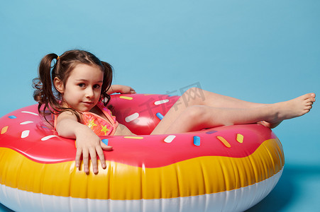 穿着泳衣的可爱小女孩，躺在蓝色背景下的充气甜甜圈游泳圈上看着相机