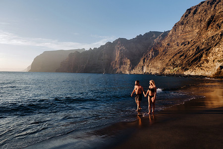 西班牙加那利群岛特内里费岛日落时分，一对热恋中的情侣在海滩上散步，背景是 Acantilados de Los Gigantes 山脉