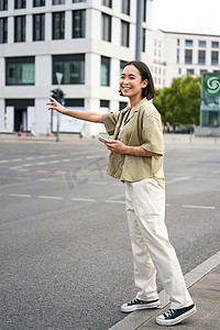 垂直拍摄的年轻女性试图赶出租车，在路上向司机挥手，手持带有汽车共享应用程序的智能手机，站在城市街道上