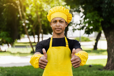 非洲黑人少年厨师戴着厨师帽竖起大拇指，身穿黄色围裙制服，为面包店做面团。