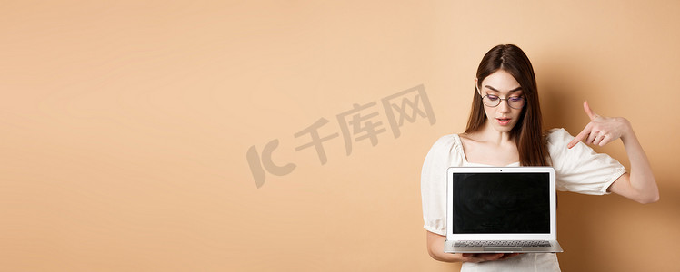 戴着眼镜的好奇女孩指着笔记本电脑屏幕，查看在线交易，在电脑上展示项目，站在米色背景中