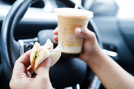 亚洲女司机拿着冰咖啡杯和三明治面包在车里吃喝，很危险，有发生事故的风险。