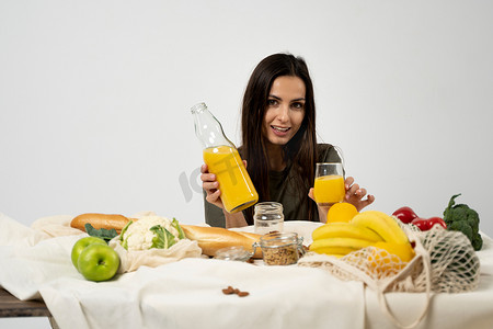身穿绿色 T 恤的女性在桌子上喝果汁，桌子上放着网眼生态袋、健康的素食蔬菜、水果、面包和小吃。
