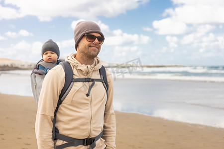 年轻的父亲在多风的沙滩上背着他的婴儿男婴儿子在背包里。