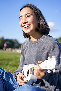 微笑的亚洲女孩唱歌和笑、弹四弦琴、学习弹奏乐器、坐在公园户外的垂直镜头