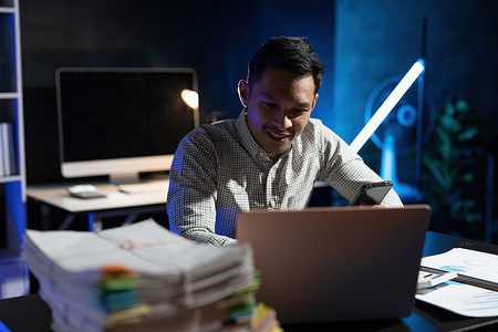 亚洲商人晚上坐在黑暗办公室的办公桌前，用笔记本电脑和手机工作。