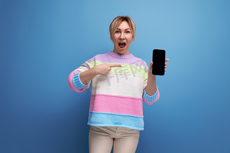身穿条纹毛衣、欢快迷人的金发年轻女性惊讶地看着智能手机的屏幕，屏幕上有蓝色背景的模型