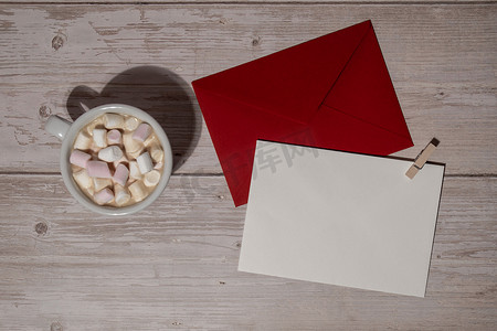 空的问候语或邀请卡模拟红色信封，白色咖啡和棉花糖在木制蓝色背景上。