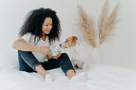 快乐的美国黑人妇女与狗共度闲暇时光，感到舒适，躺在床上摆着白色床上用品的水平镜头。