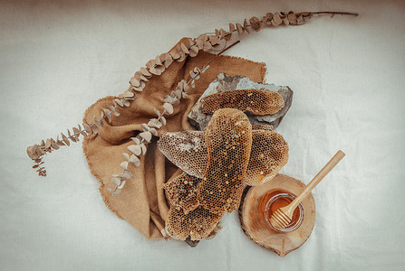 美味的蜂蜜摄影照片_美味的蜂蜜从玻璃罐上的新鲜蜂窝中滴落下来，木制蜂蜜勺子放在麻布上。