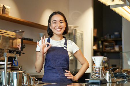 微笑的韩国咖啡师的画像，柜台的女孩，穿着蓝色围裙，在咖啡店工作，展示了好的标志