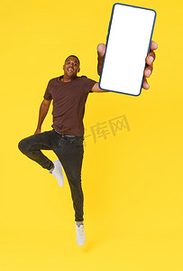 手机线上广告摄影照片_精力充沛的非洲裔美国学生被拍到在黄色背景上手持手机跳跃，手机上有空白屏幕。