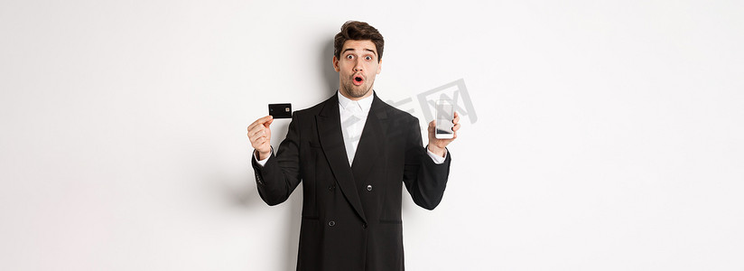 穿着黑色西装的英俊商人的形象，看起来很惊讶，并用手机屏幕展示信用卡，站在白色背景下