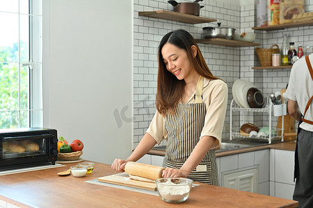 微笑的家庭主妇穿着围裙，在餐桌上用针卷面团，在现代厨房里做面包