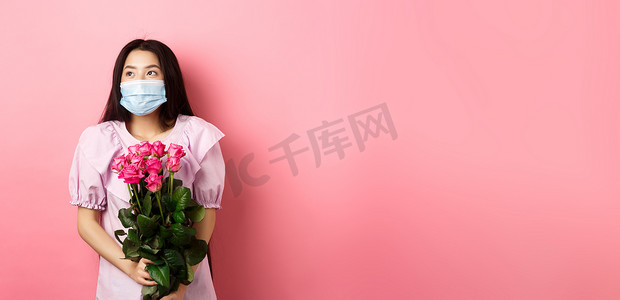 戴着医用面具的浪漫亚洲女孩用梦幻般的眼睛望着空荡荡的空间，在情人节拿着一束玫瑰，在大流行病期间与情人约会