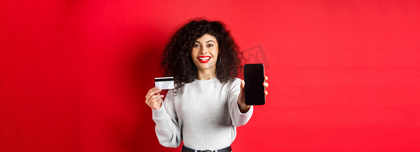 展示塑料信用卡和空智能手机屏幕的快乐漂亮女孩，展示账户或应用程序，站在红色背景上