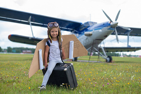坐飞机的儿童摄影照片_一个穿着飞行员服装的小女孩坐在机场的复古手提箱上等待航班起飞。
