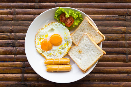 炒鸡蛋、香肠、新鲜西红柿、绿色沙拉和烤面包，放在棕色竹桌上的白盘子里。