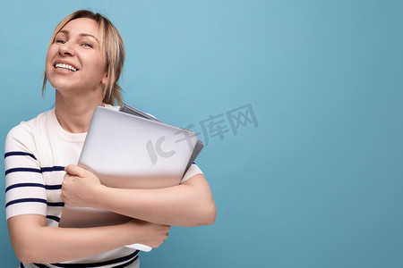 条纹毛衣摄影照片_蓝色背景中一位自信的金发女性自由职业者的肖像照片，她身穿条纹毛衣，手里拿着一台笔记本电脑