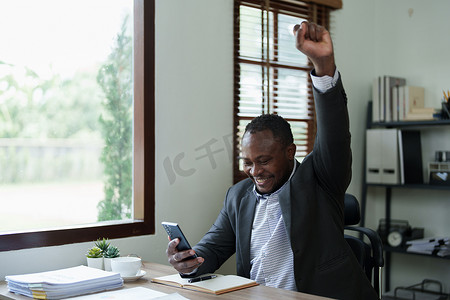 中年男子美国非洲商人手持智能手机，双手举起赢家，这是一种姿态，很高兴成功庆祝成就成功