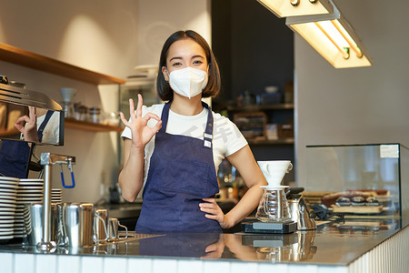 冠状病毒表现摄影照片_亚洲女咖啡师，戴医用面具的女孩，表现不错，在柜台后的咖啡馆工作，煮咖啡，在大流行病期间与客户合作